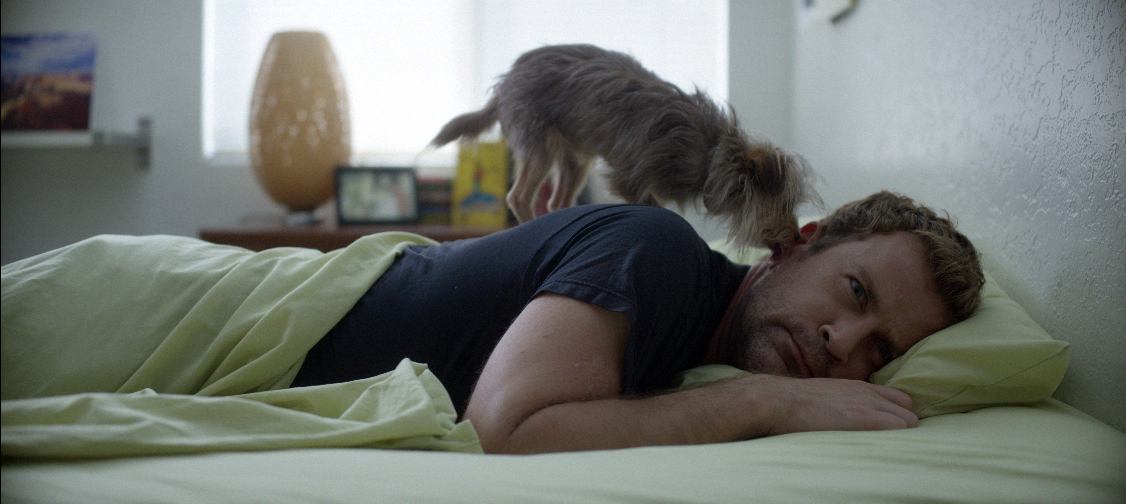 Yorkie Terrier licking the ear of a white, blue-eyed man who is laying on his bed