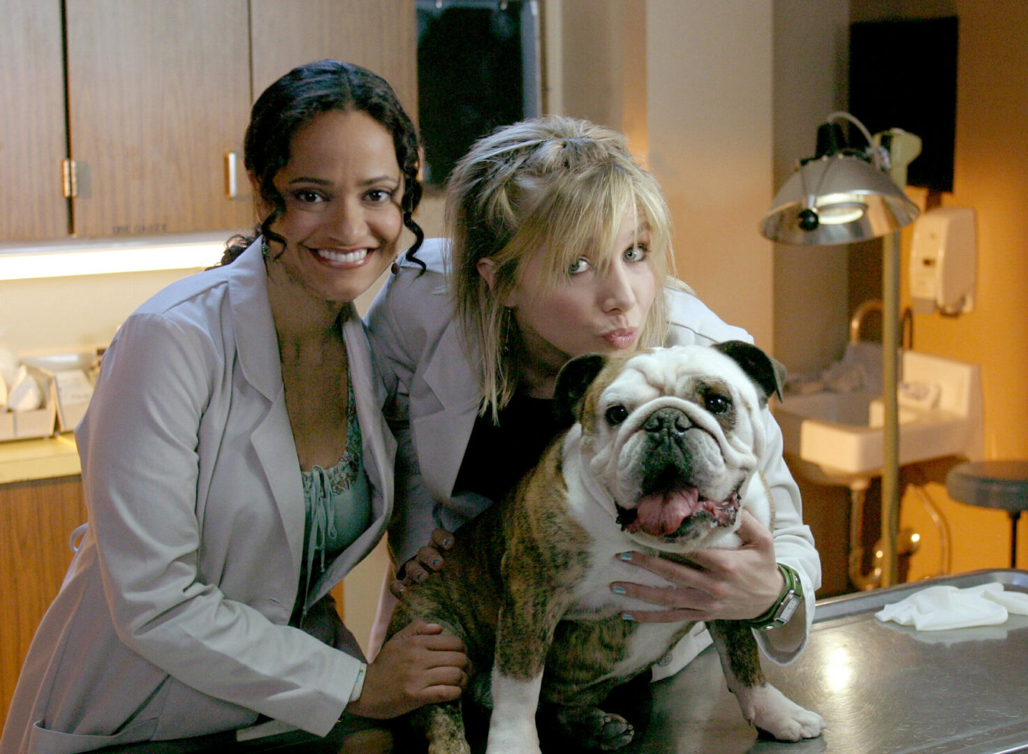 Two actresses in lab coats posing with a panting, brown-and-white bulldog --