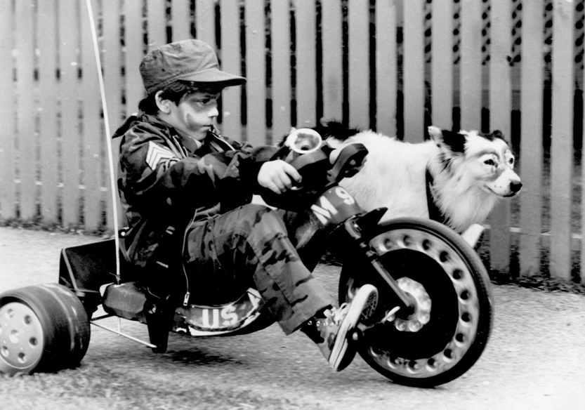 Black-and-white picture of a boy riding a toy motorbike with a Border Collie by his side