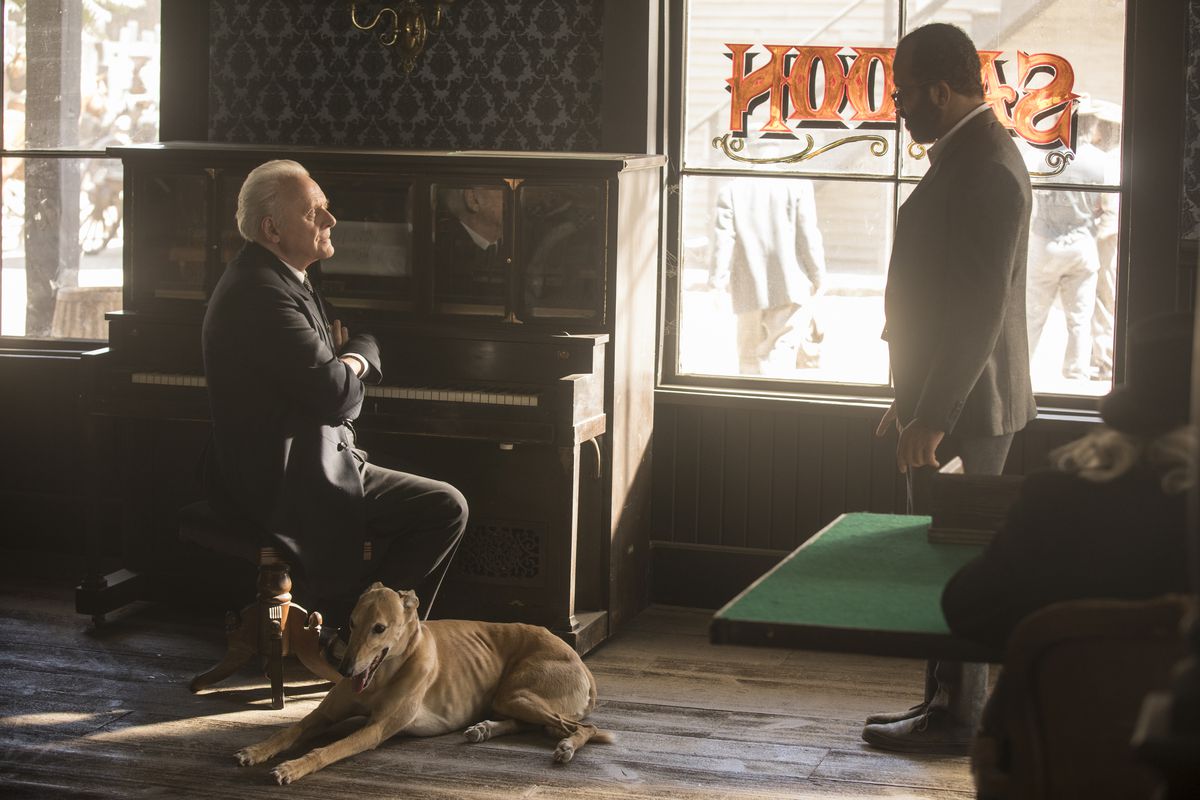 Light brown Greyhound sitting on the floor next to a white guy in a saloon scene in Westworld